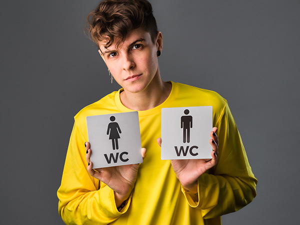 Image of a person in a yellow shirt holding two gendered bathroom signs.