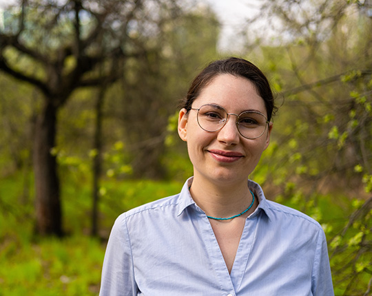 A photo of a woman with glasses looking at the camera with trees in the background.