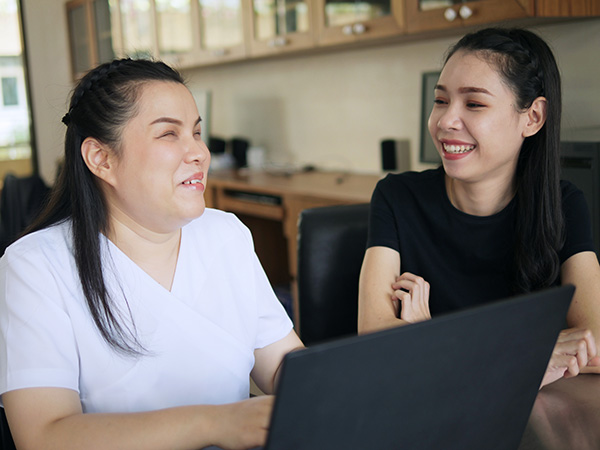 Asian women co-workers smile in workplace. One woman is a person with blindness disability using a laptop computer with a screen reader program for visually impaired people.