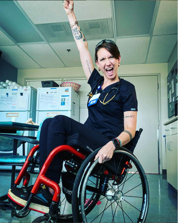 Ryann Mason in black scrubs sits in a wheelchair and pumps fist with an excited expression on her face.