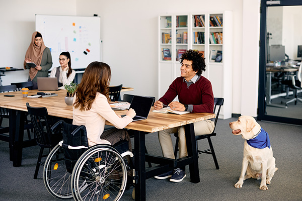 A man with a canine service dog meets with a woman using a wheelchair in a diverse office.  