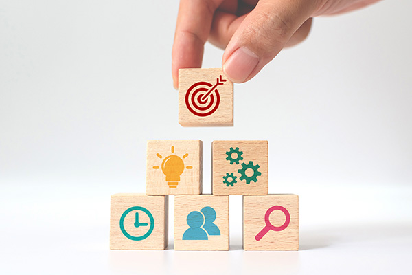 Hand placing six wooden cube blocks in a stack on a white background. Each block has different icons including a clock, people, magnifying glass, light bulb, gears, and a target.