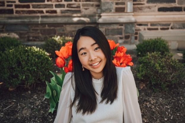 A Korean American woman wearing a white shirt with orangish red flowers and other plants in the background.