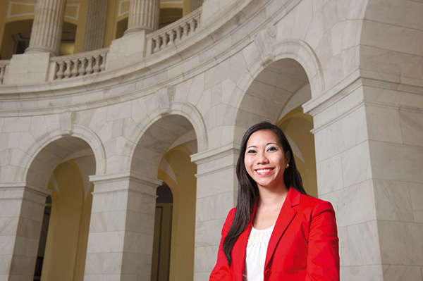 Headshot of smiling Krystal Ka’ai in a red blazer standing inside a building with grand white stone domed doorways and pillars