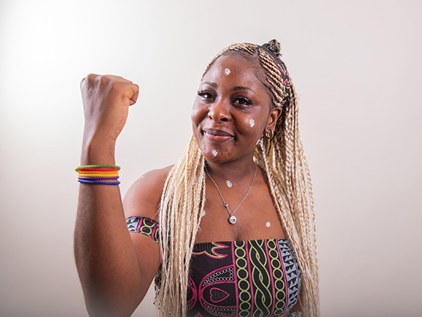 A Black lesbian woman with her arm raised shows her biceps and wears colorful, rainbow colored bracelets. She wears an African print halter top with black, green, purple, and pink geometric shapes.