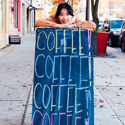 Olive Jung smiles and stands outside on a sidewalk as she rests her arms and head on a tall chalkboard that reads, Coffee! Coffee! Coffee! Coffee!