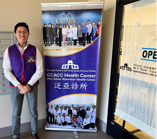 Author and IRTA Fellow, Simon Liu wearing a blue vest and standing in front of a Chinese Culture and Community Service Center banner. 