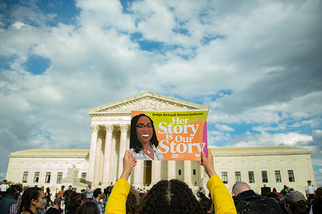 Person facing away from camera faces the court in DC and holds up colorful poster with Judge Ketanji Brown Jackson and the words ‘Her Story is Our Story ‘