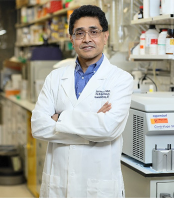 Bangladeshi American Deputy Chief and Senior Investigator, Dr. Javed Khan in his lab, wearing a white lab coat, standing with his arms folded in front of him.