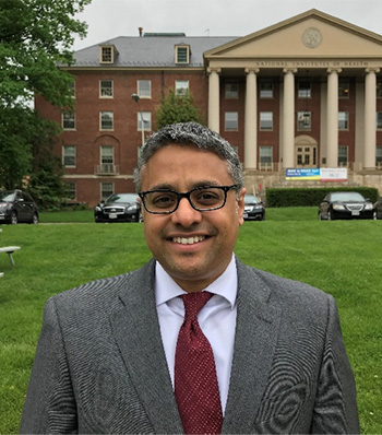Indian-American Science Policy Branch Chief Sandeep Dayal, Ph.D., standing in front of an NIH campus building, several cars, and a grassy area.