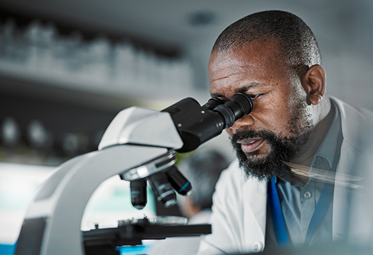 Black male scientist peers through a microscope in a lab.