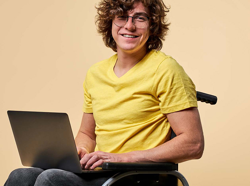 Smiling man sittin in a wheelchair with a laptop on his lap.
