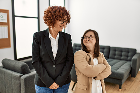An older woman and a woman with down syndrome work and smile at each other in the office.