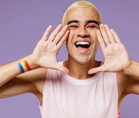 Image of an individual smiling and yelling with a pride flag tattoo standing in front of a light purple background.