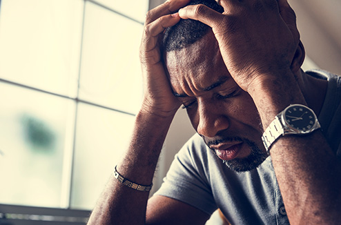 A stressed looking Black man furrowing his brow and closing his eyes with both hands grasping his head.
