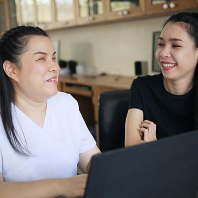 Asian women co-workers smile in workplace. One woman is a person with blindness disability using a laptop computer with a screen reader program for visually impaired people.