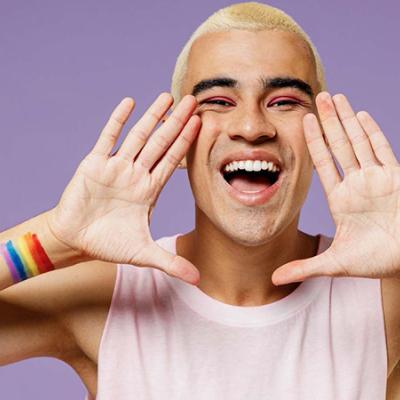 Image of an individual smiling and yelling with a pride flag tattoo standing in front of a light purple background. 