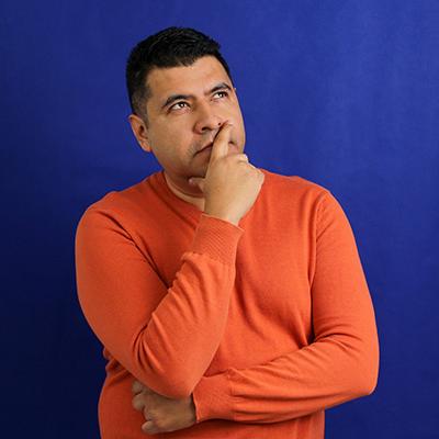 A curious Hispanic/Latino man stares with his hand over his mouth stands in front of a blue background. 