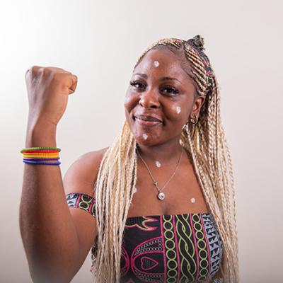 A Black lesbian woman with her arm raised shows her biceps and wears colorful, rainbow colored bracelets. She wears an African print halter top with black, green, purple, and pink geometric shapes.