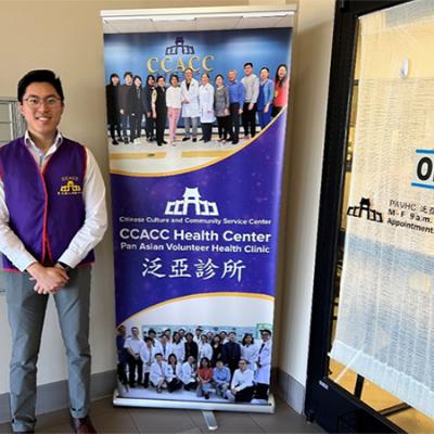 Author and IRTA Fellow, Simon Liu wearing a blue vest and standing in front of a Chinese Culture and Community Service Center banner. 